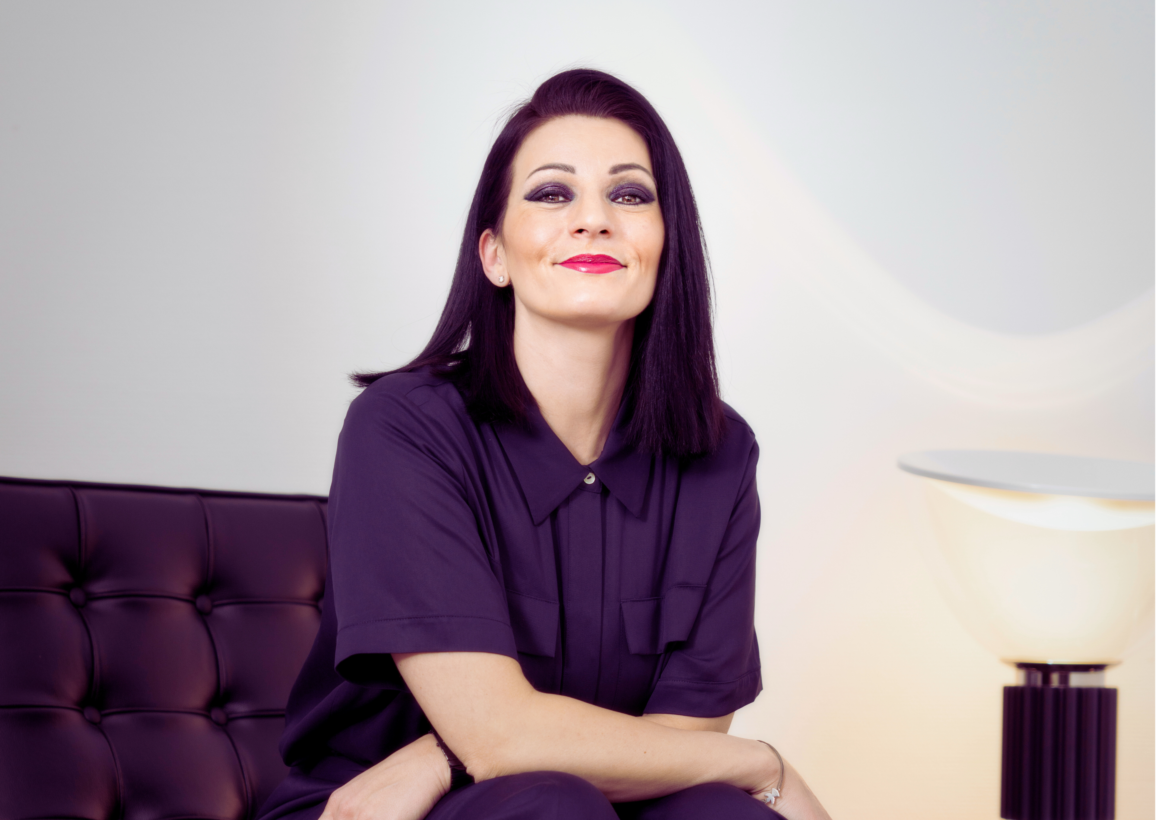 Eine lächelnde Frau in einem dunklen Outfit sitzt selbstbewusst auf einem Sofa, passend zum Thema Finanzen für Geschäftsführer.